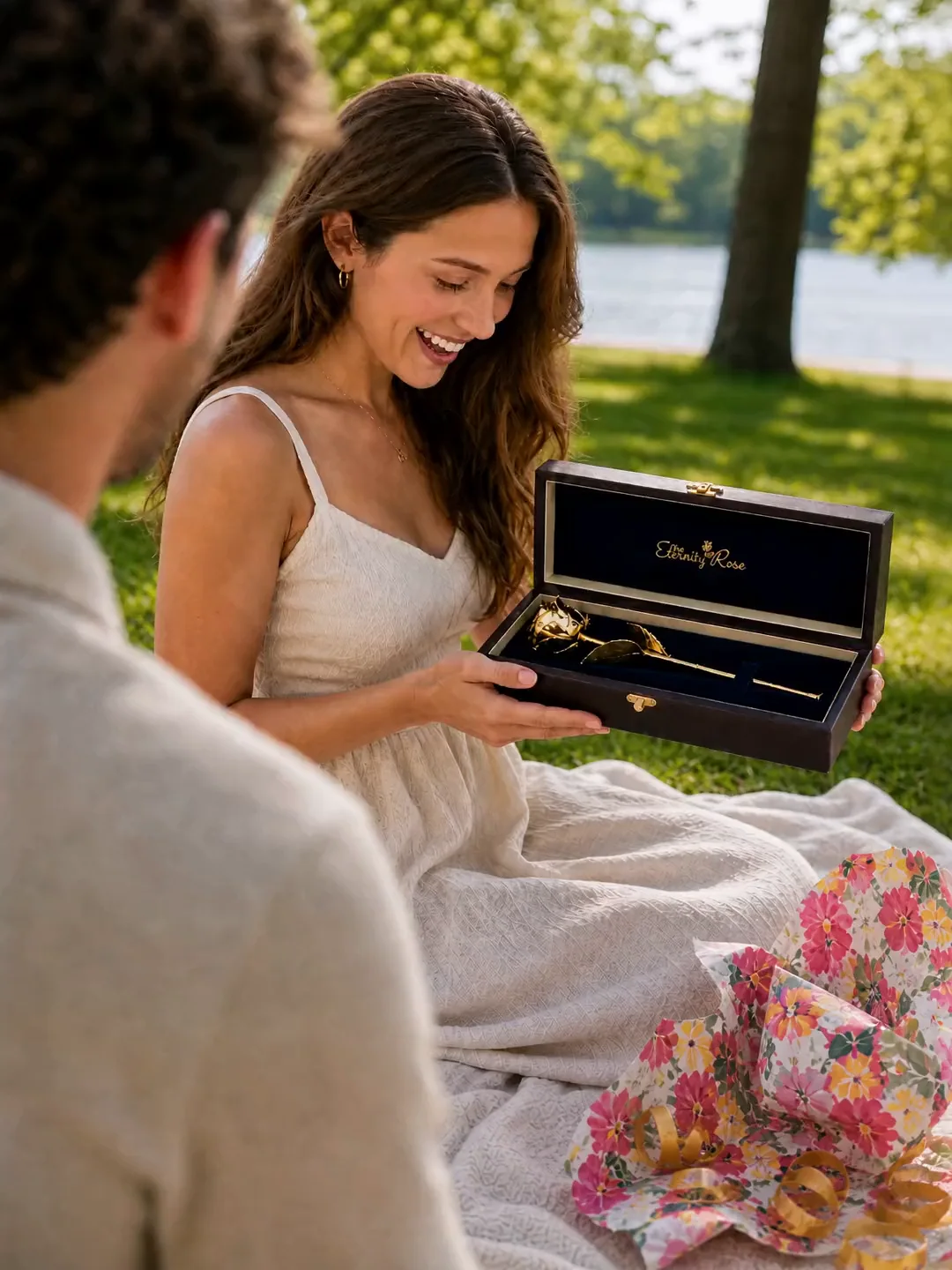 A woman opening a gold Eternity Rose presentation case at a lakeside picnic while her partner looks on